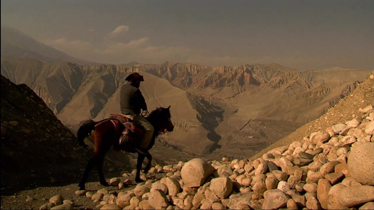 Travelers ride horses along a rugged mountain trail in the high altitude landscape of the Himalayas. The scene captures the vast, arid terrain and remote scenery of a mountain plateau.