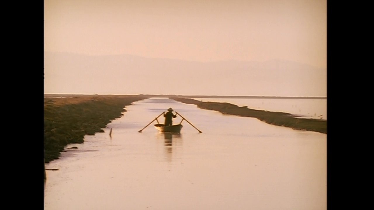 A man drives a red pickup truck along a remote waterside path before transitioning to scenes of rowing a boat on a calm lake and resting in quiet contemplation. The sequence captures a narrative of travel, introspection, and peaceful outdoor activity.