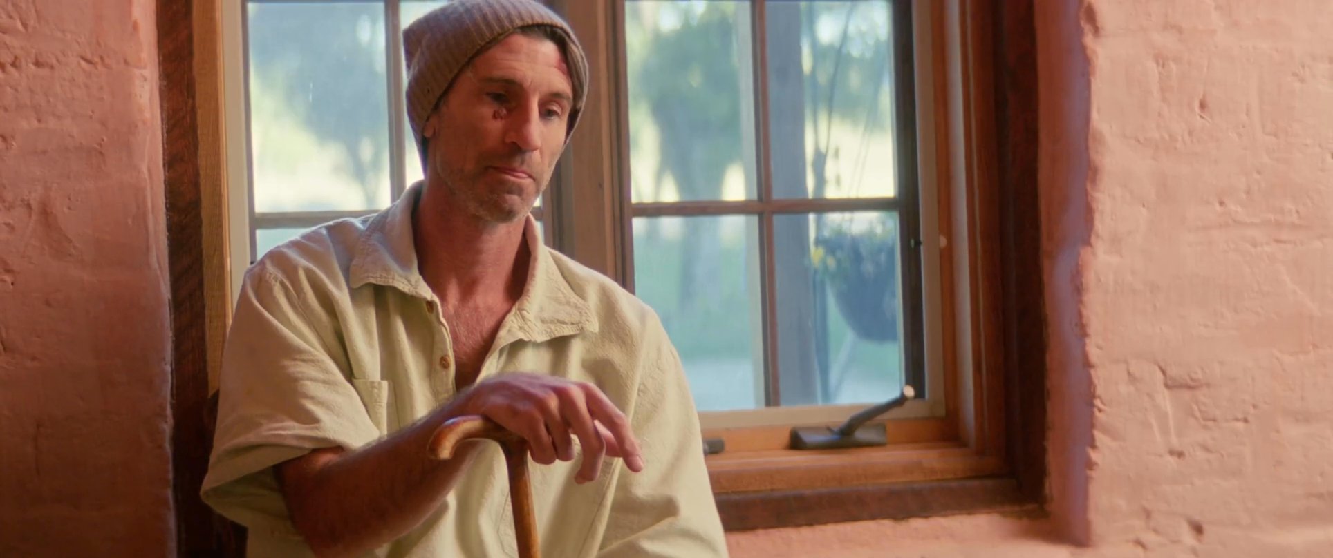 Two men engage in a focused conversation within a sunlit, textured interior space. One man rests on a cane while the other observes him during their exchange.