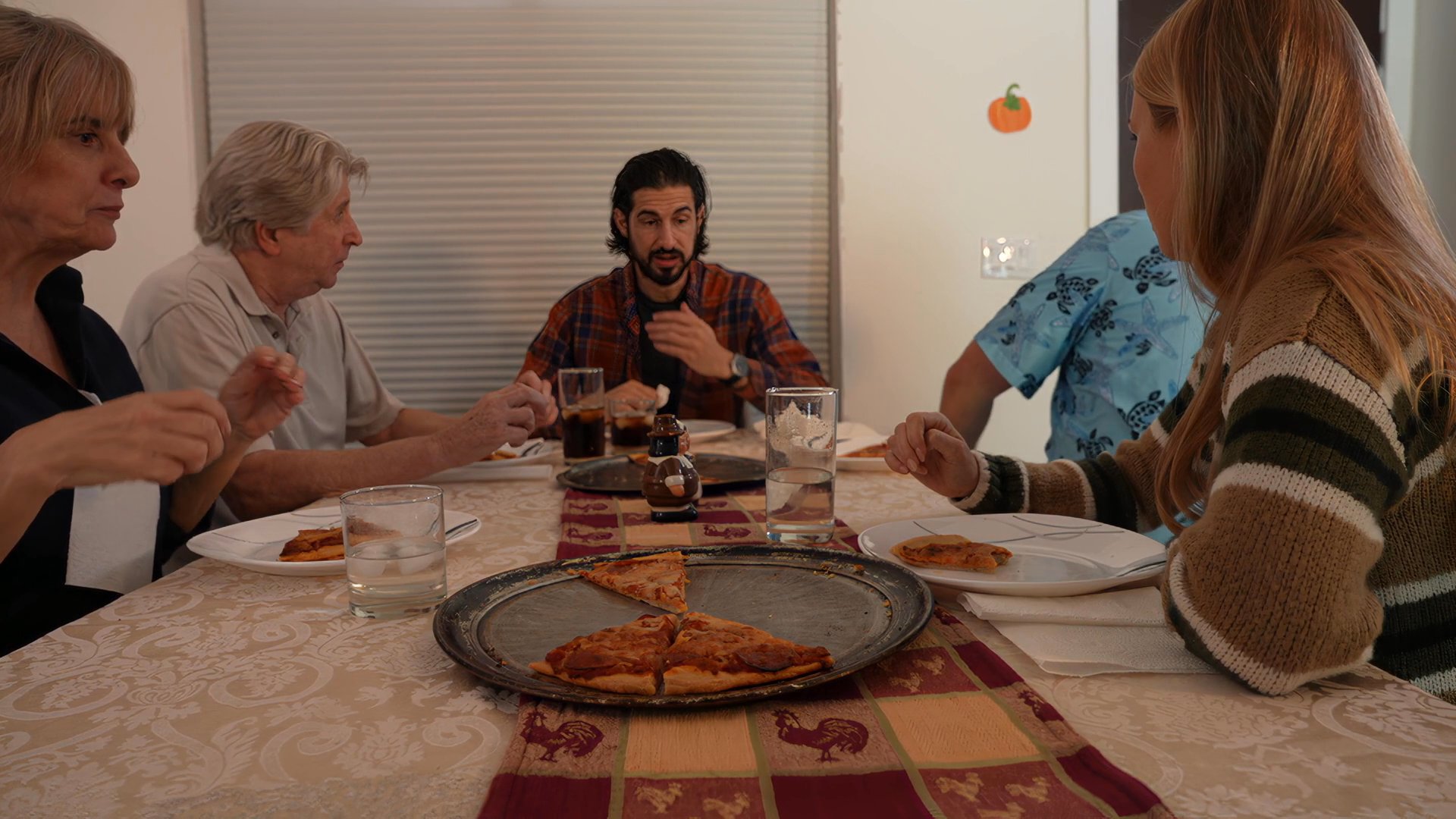 A family shares a meal together at a dining table decorated for the Thanksgiving holiday. The group engages in conversation and eats while gathered in a residential dining room.