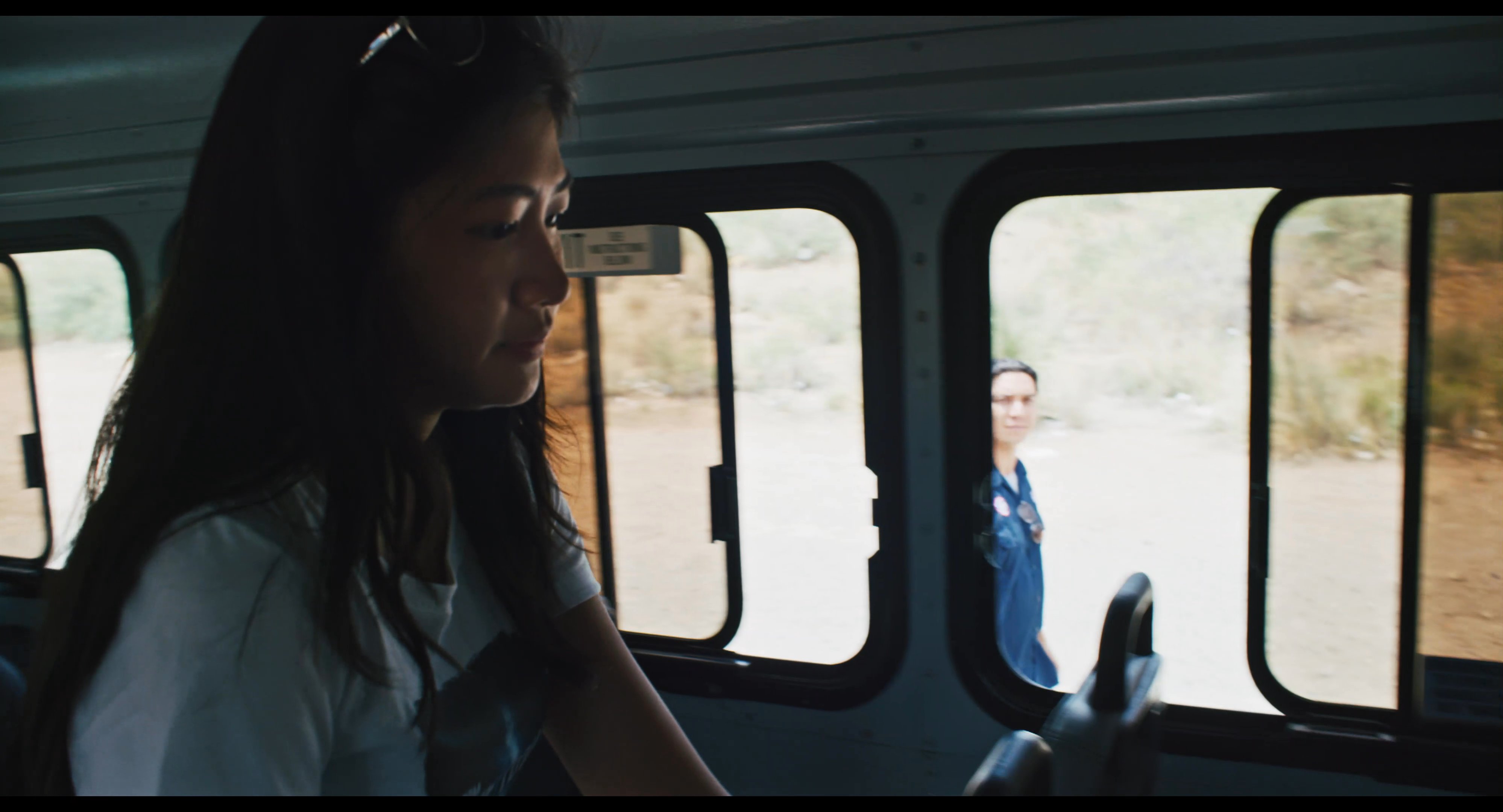 A passenger boards a bus while the driver observes before the vehicle pulls away. The driver waves goodbye as the passenger watches from inside, marking the start of a journey.