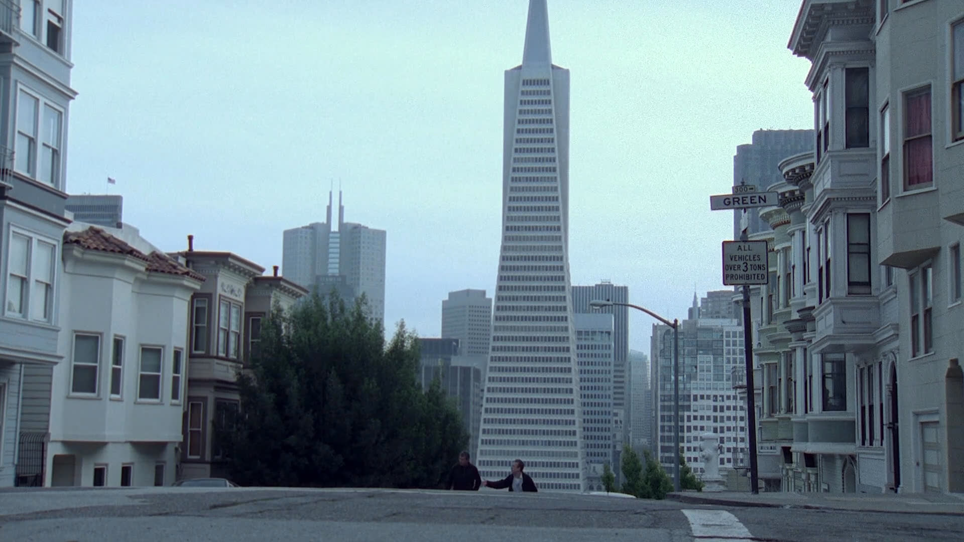 A man observes a tense encounter in an interior space before the scene shifts to a wide, establishing view of a street in San Francisco with the Transamerica Pyramid in the background. Pedestrians and a cyclist navigate the city street under an overcast sky.