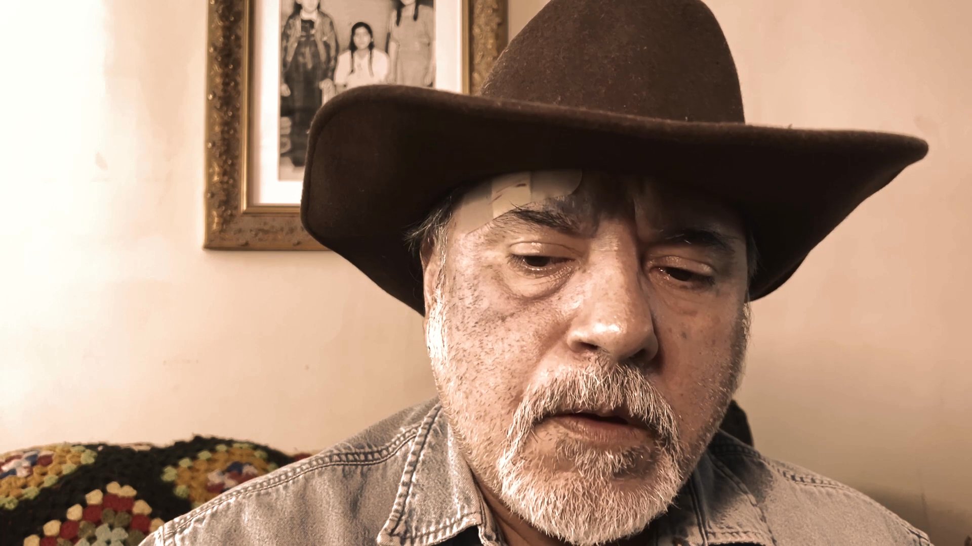 A man wearing a western-style cowboy hat delivers a direct-to-camera monologue in an indoor residential setting.