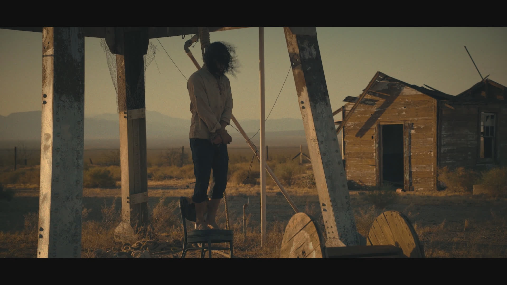 A long-haired man stands alone beside a metal structure in a remote, sun-baked settlement, with a small shack and power lines in the distance. He sighs and speaks briefly, his face tense and troubled.