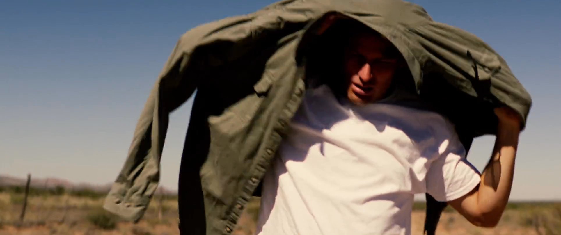 A man stands in an arid desert landscape and puts on a green utility jacket while wearing sunglasses. The bright sunlight emphasizes the heat of the environment as he prepares for his journey.