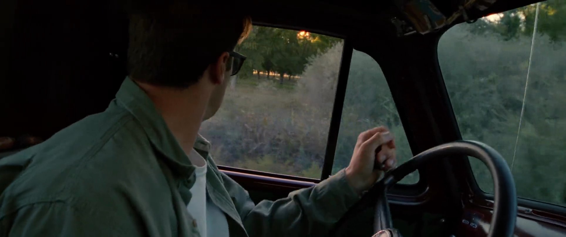 A man drives a vintage truck down a dirt road during golden hour while consulting a map to navigate the rural landscape.