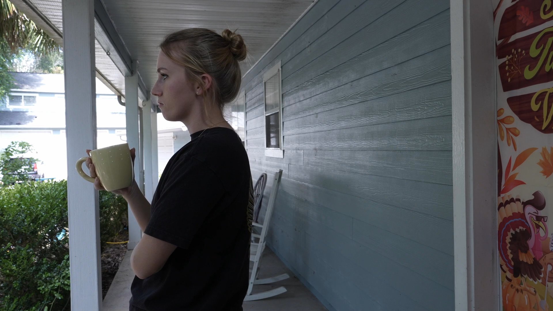 A woman stands on a porch holding a coffee mug, appearing deep in thought before expressing a sudden, intense emotional outburst.