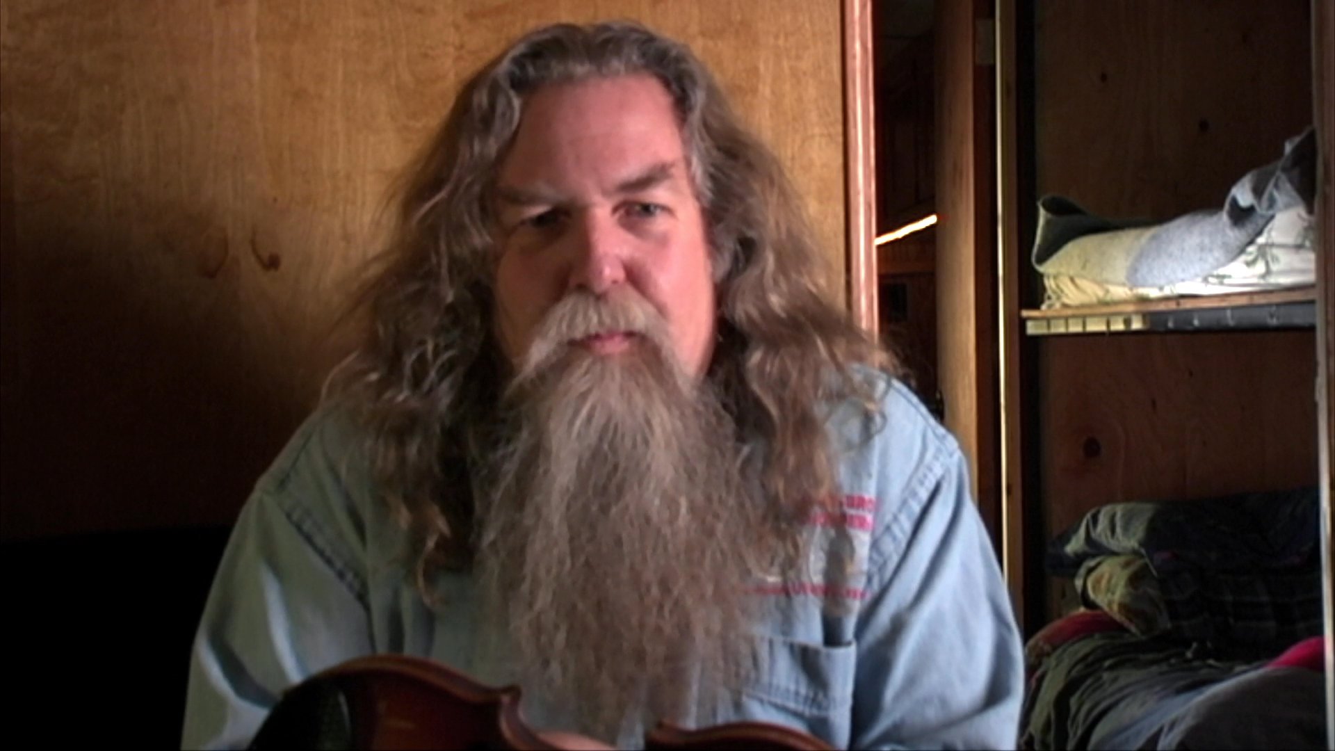 A long-haired, bearded musician engages in a lively storytelling performance while holding a violin. He gestures expressively to illustrate his narrative against a wood-paneled background.
