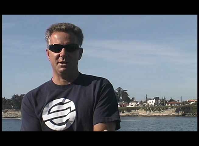 A surfer rides a wave in the ocean before transitioning to an interview with a man standing along a coastal landscape. The footage highlights aquatic activities paired with professional commentary.
