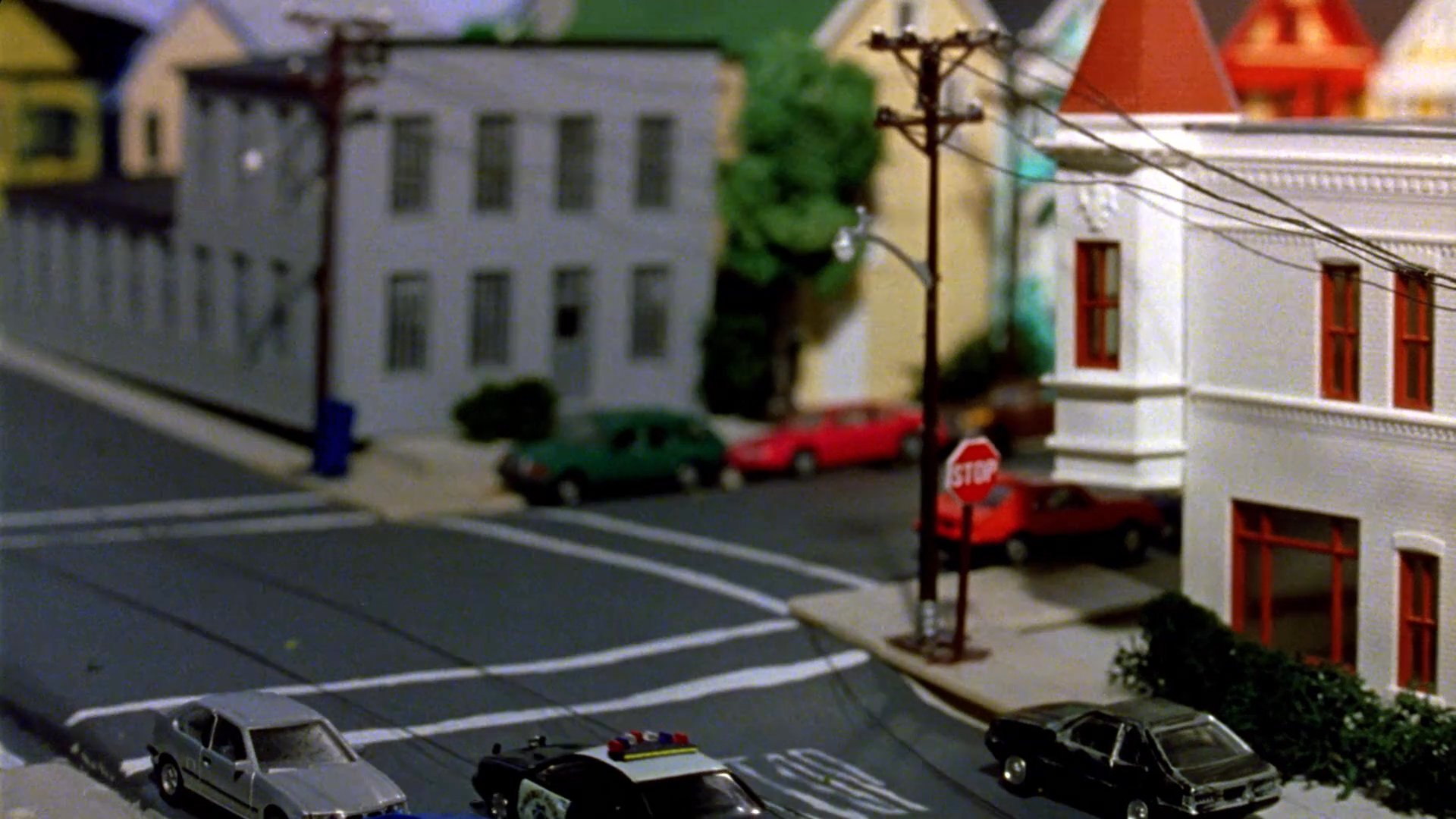 Passengers in a vehicle watch a miniature diorama of a suburban street as toy cars move through a town intersection.