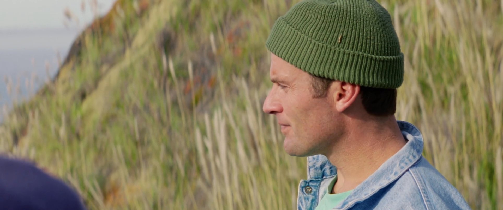 Two men engage in a conversation while standing along a grassy coastal cliffside.