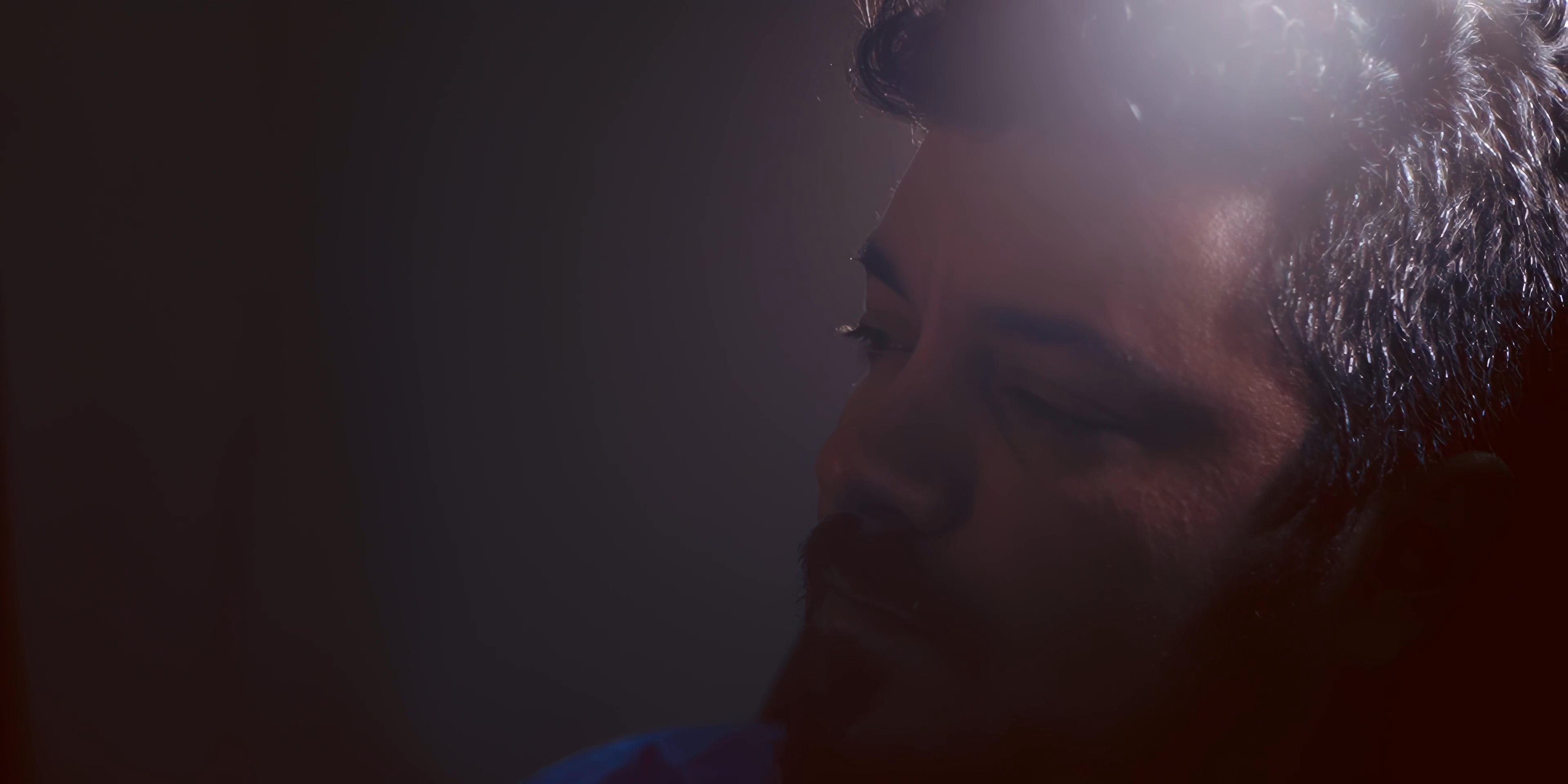 A man sits alone in a dimly lit room, holding a glass and bottle of alcohol before covering his face in a gesture of profound distress.