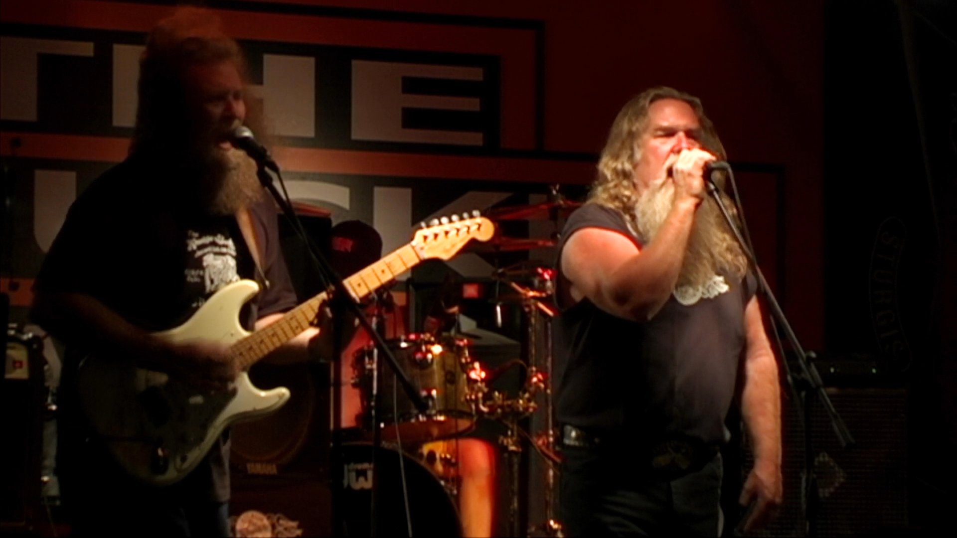 A long-haired, bearded vocalist performs on a dimly lit stage accompanied by a guitarist. The band plays a rock set before shifting focus to the instrumentalists.