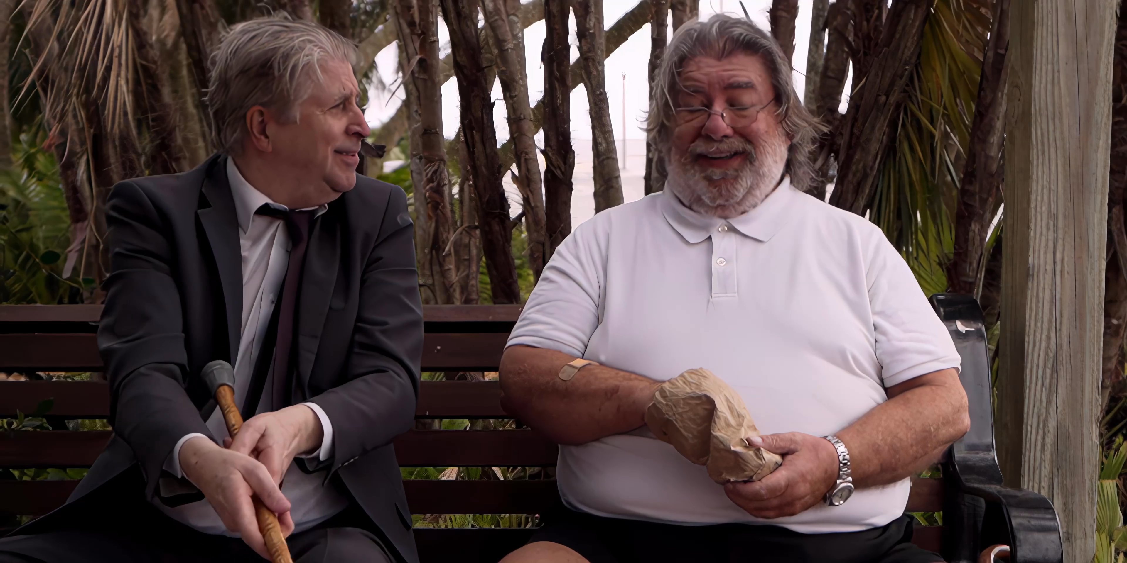 Two men engage in an animated discussion while seated together on a park bench surrounded by trees. They gesture expressively as they converse in a relaxed, informal setting.