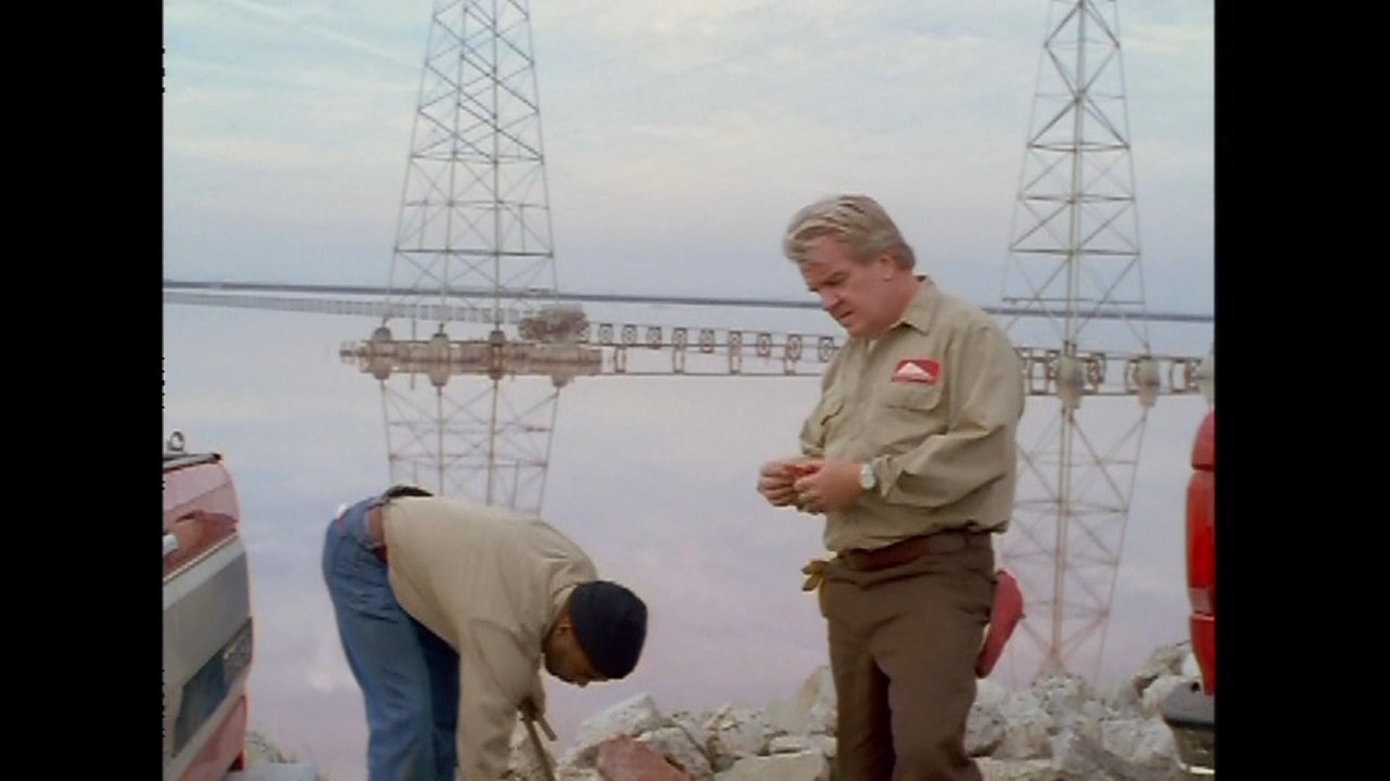 Two workers in uniform systematically gather materials from a rocky landscape and load them into the bed of a pickup truck. The activity takes place at a remote industrial location marked by tall support towers.