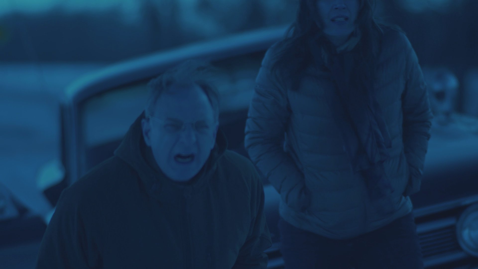 A man and a woman engage in an intense argument by their vehicle in a desolate, snow-covered landscape at dusk.