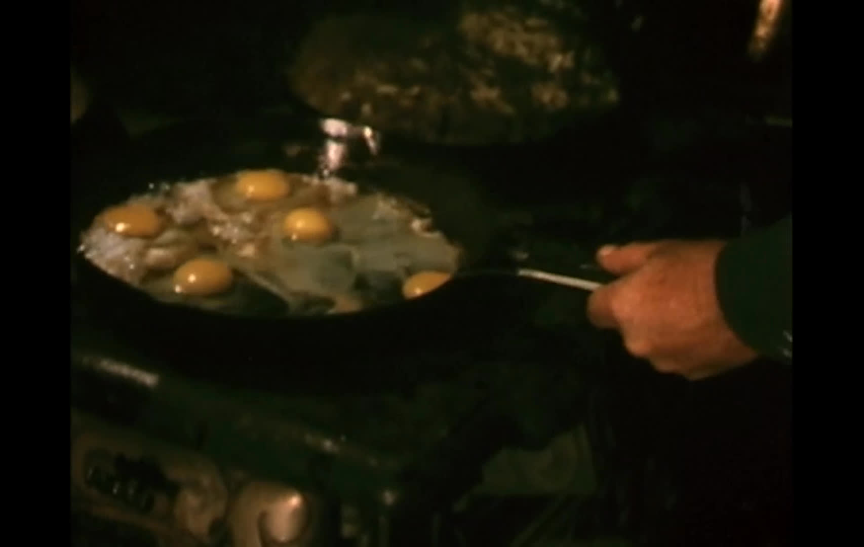 A rustic cabin and glowing wood stove set the scene as a cook tends skillets of sizzling eggs and bacon. Cowboy hats hanging above the stove reinforce the ranch-life atmosphere.
