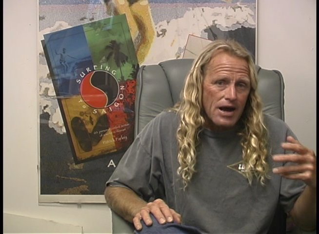 A man with long blonde hair sits in a chair while gesturing and speaking during an interview in front of a surfing-themed poster.