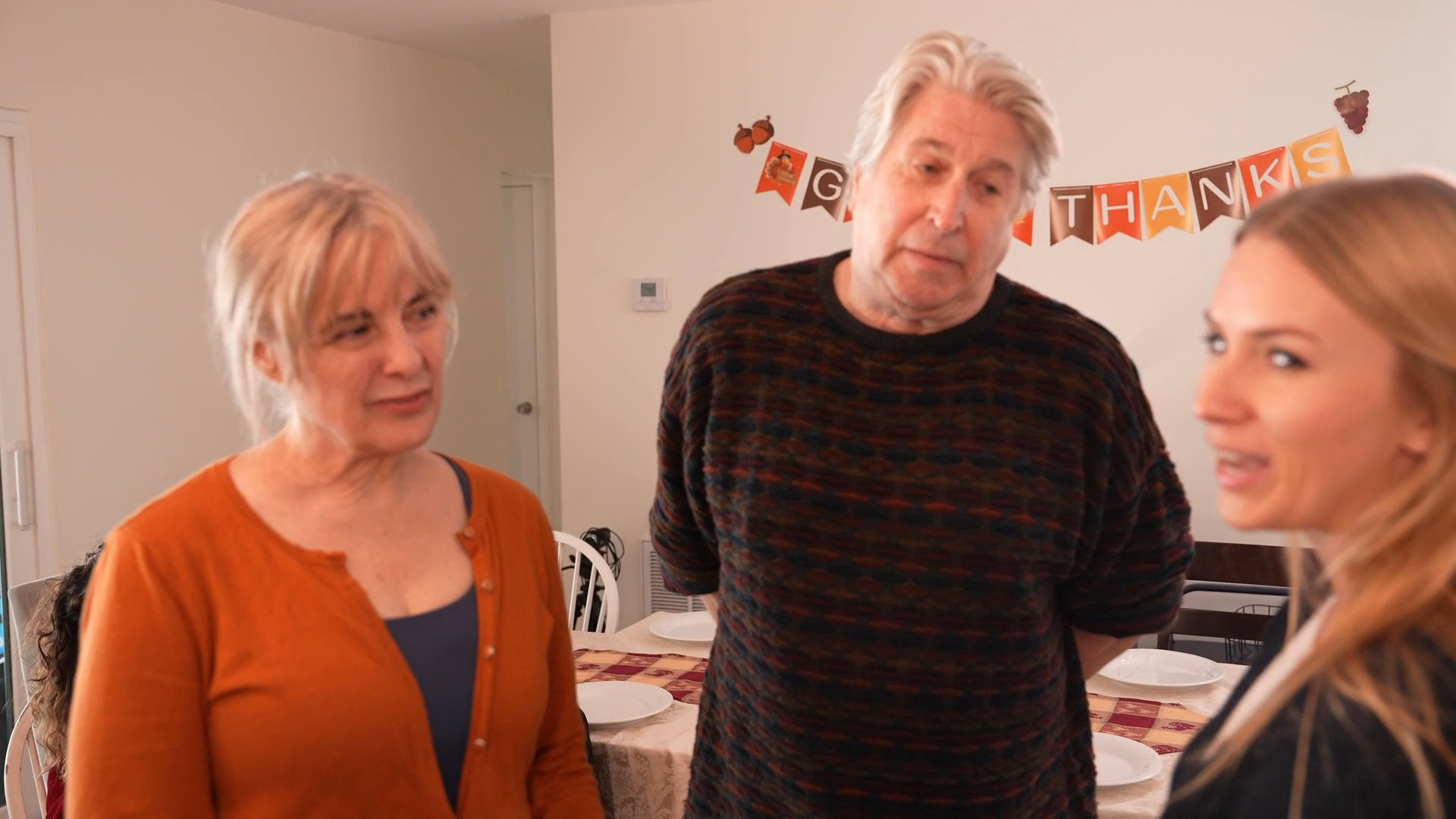A group of adults gather in a home decorated for Thanksgiving, engaging in conversation and welcoming guests at the entrance.