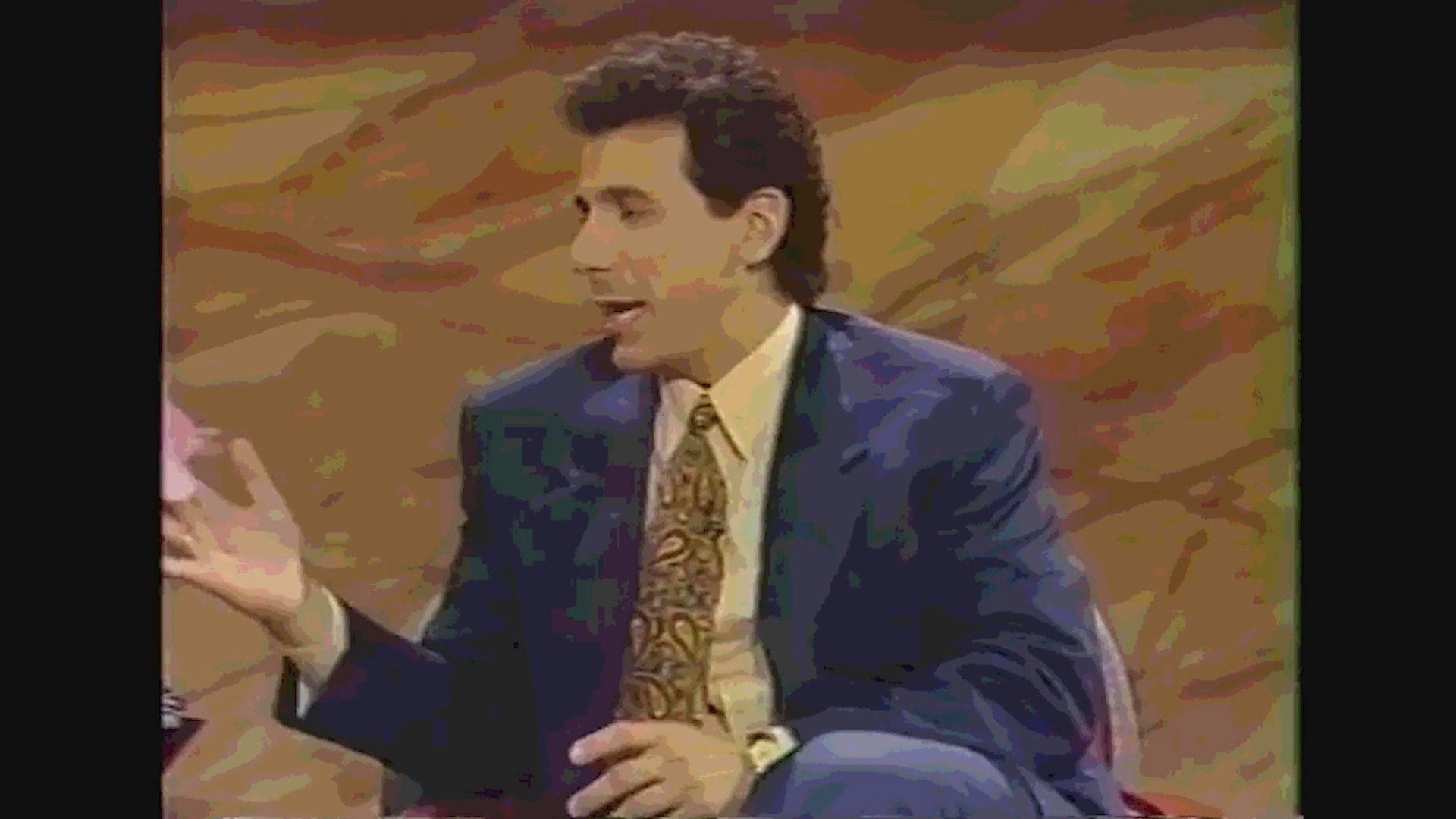 A television host interviews two male guests on a colorful studio set, facilitating a casual and engaging conversation.