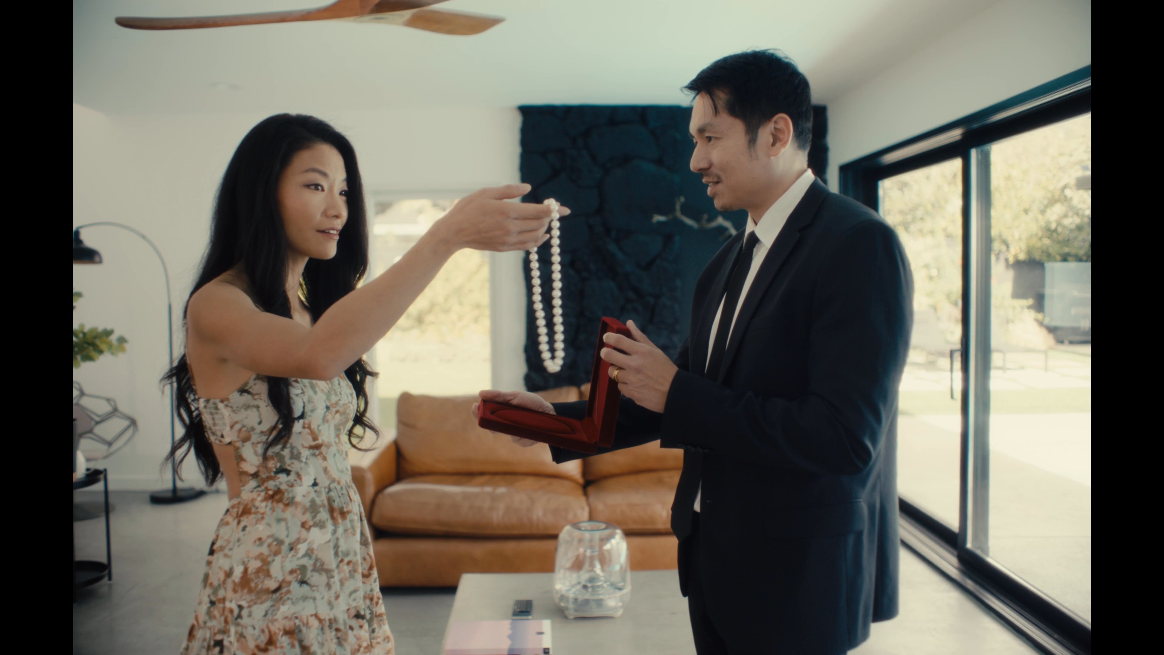 A man presents a jewelry box containing a pearl necklace to a woman within a contemporary living space. The woman receives the piece, admiring the gift as the man observes.