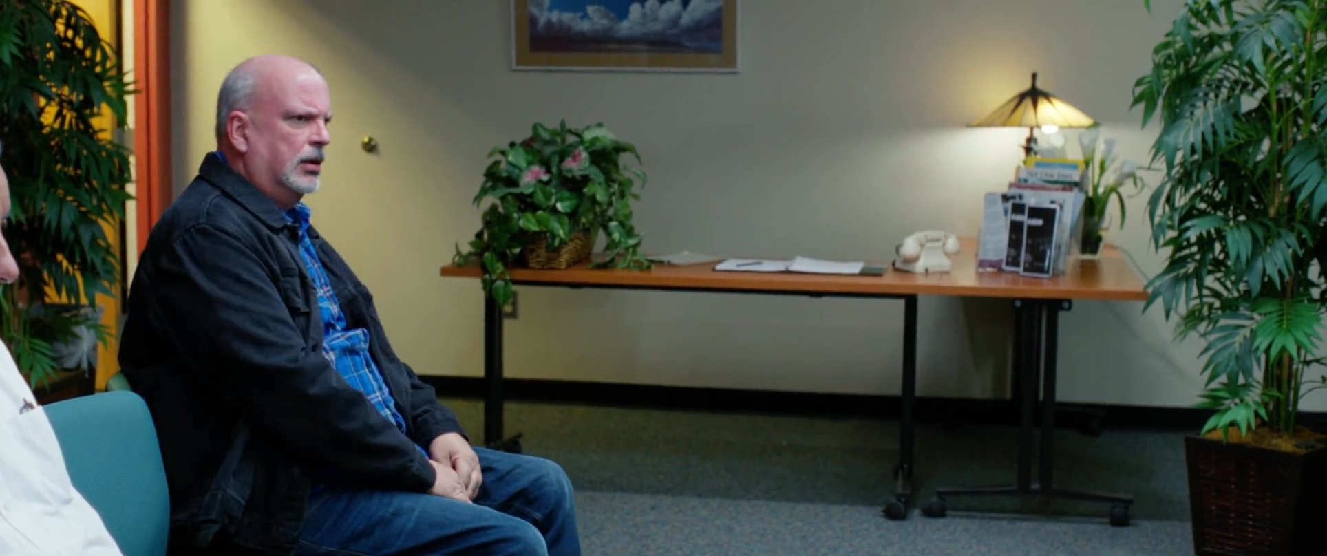 Two men conclude a conversation in a waiting area before walking outside to board a vehicle together, leaving a third man seated behind.