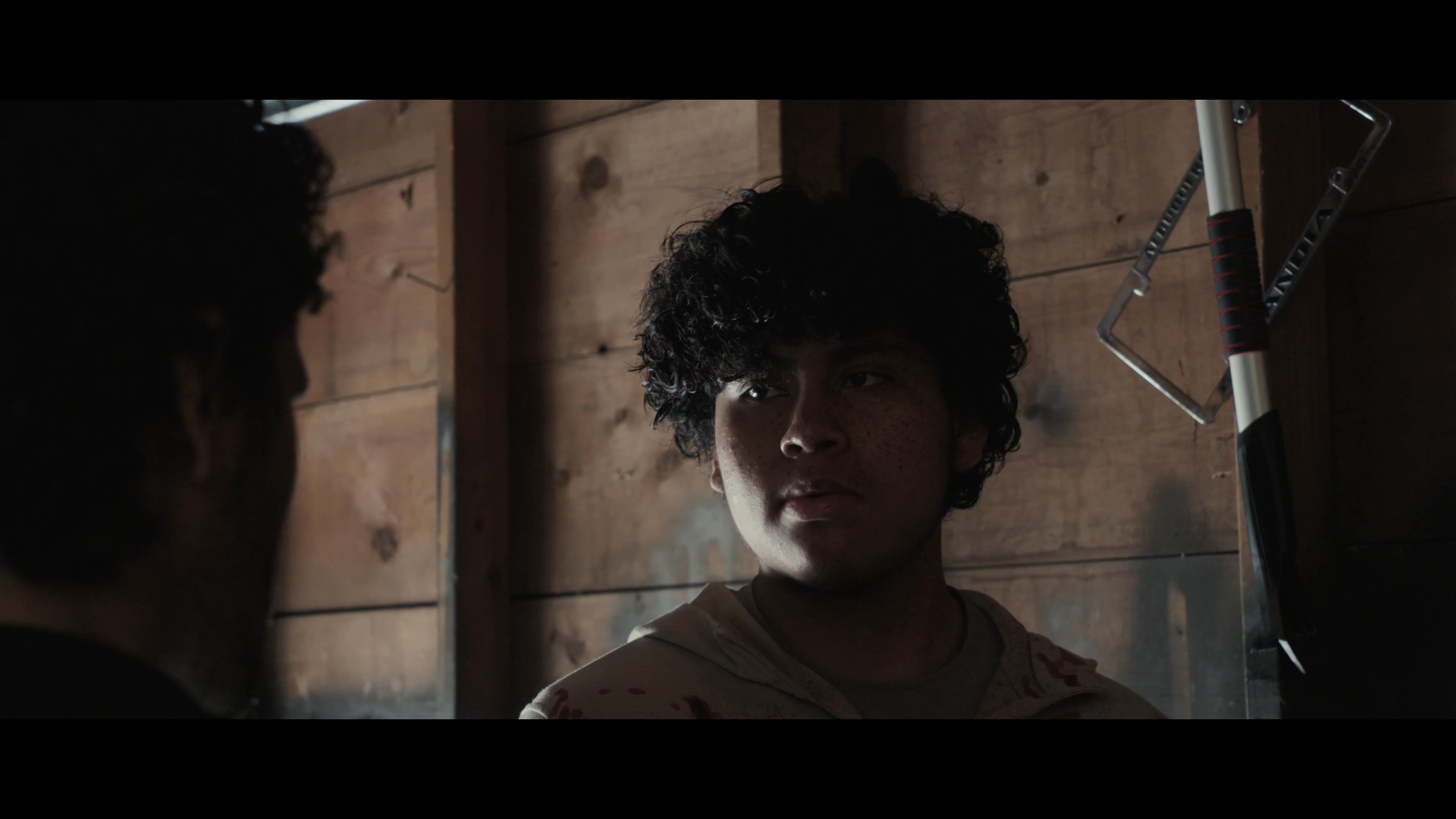 Two men engage in an intense, serious conversation inside a dimly lit garage. The atmosphere remains heavy with unspoken tension as they exchange words.