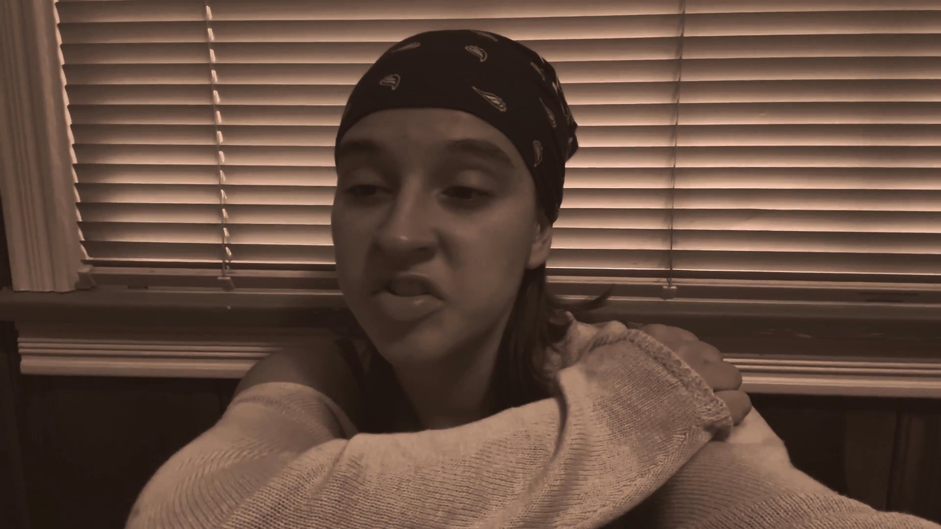 A woman speaks directly to the camera with a serious demeanor while seated indoors against a window blind background. The scene shifts briefly to an older man, suggesting a multi-part interview or interactive dialogue.
