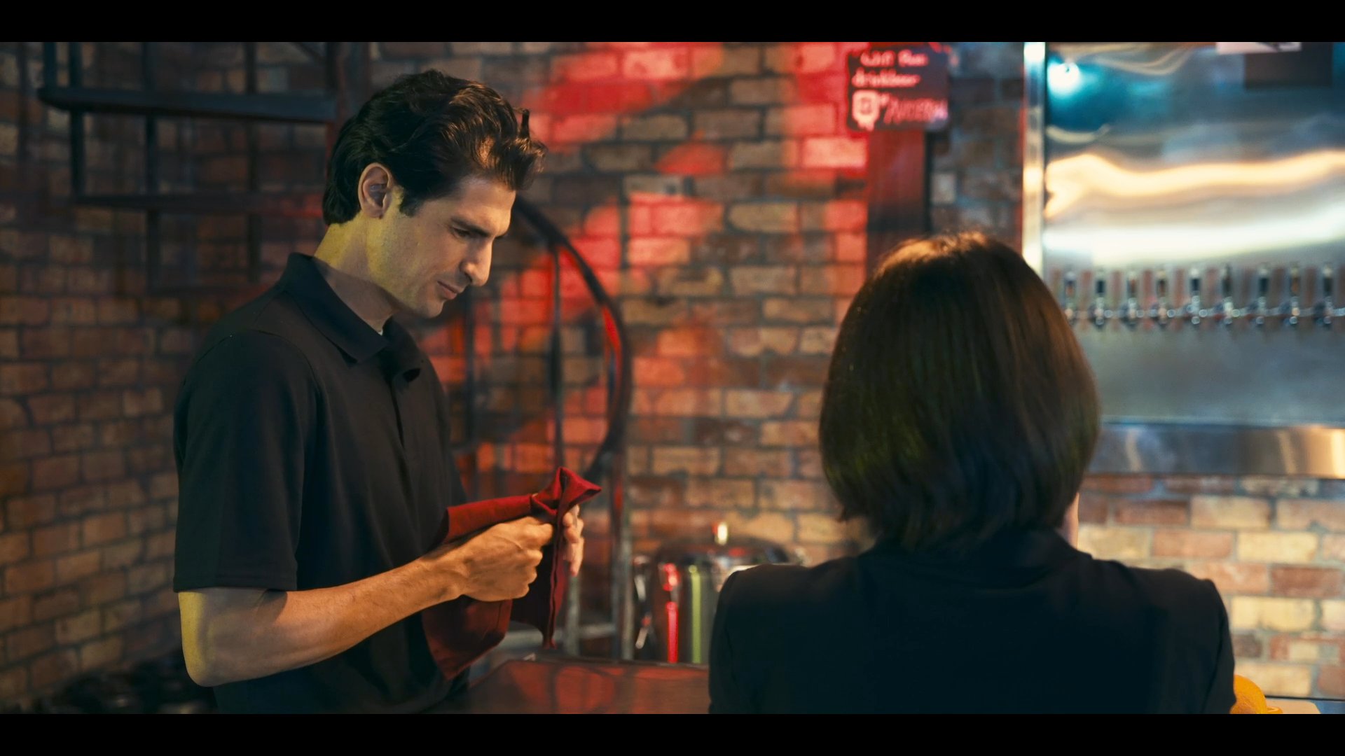 Staff members discuss operations within a brewery setting before one attendant assists a customer at the bar.