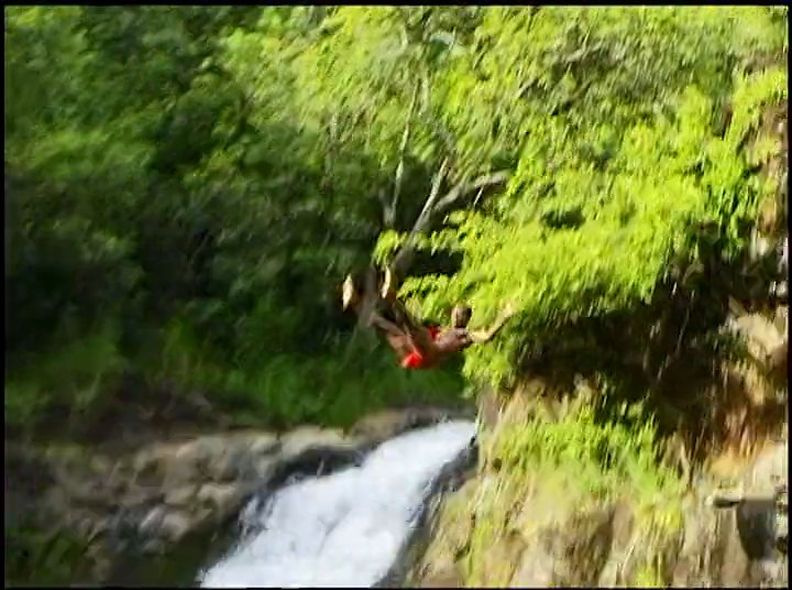 Athletes transition from rowing on open water to recreational cliff jumping at a scenic waterfall. The sequence captures individuals leaping into a natural pool surrounded by dense forest foliage.