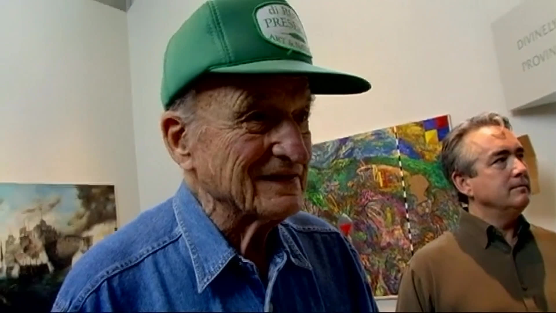 An older man in a green cap discusses his affection for a colorful, whimsical assemblage sculpture in a gallery, alongside another visitor. The view shifts to two men carefully handling a large object in a work area.