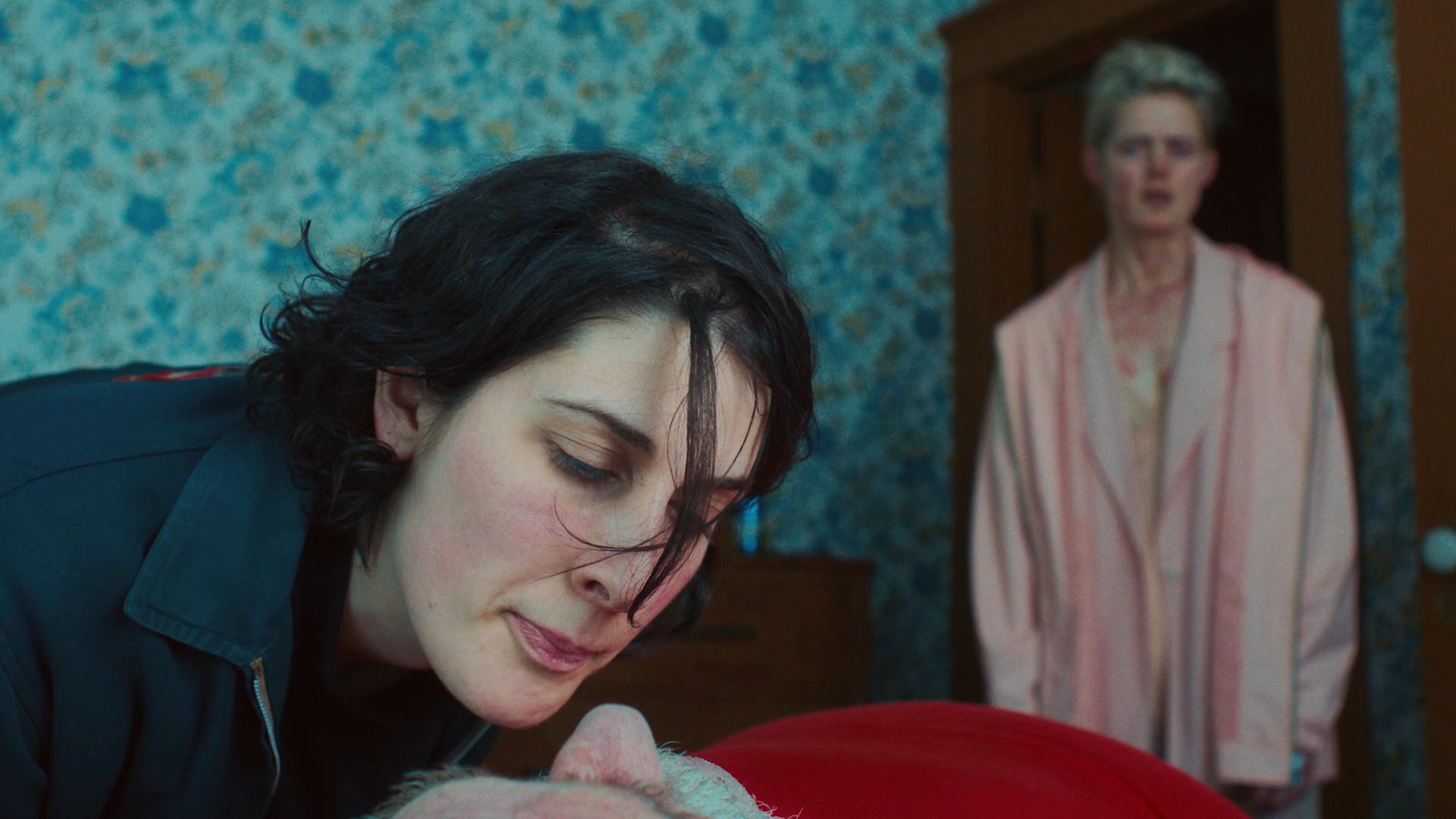 Two individuals interact in a confined bedroom space, alternating between periods of close caregiving and intense interpersonal observation. The atmosphere remains heavy with unspoken conflict as they attend to an elderly person resting in bed.