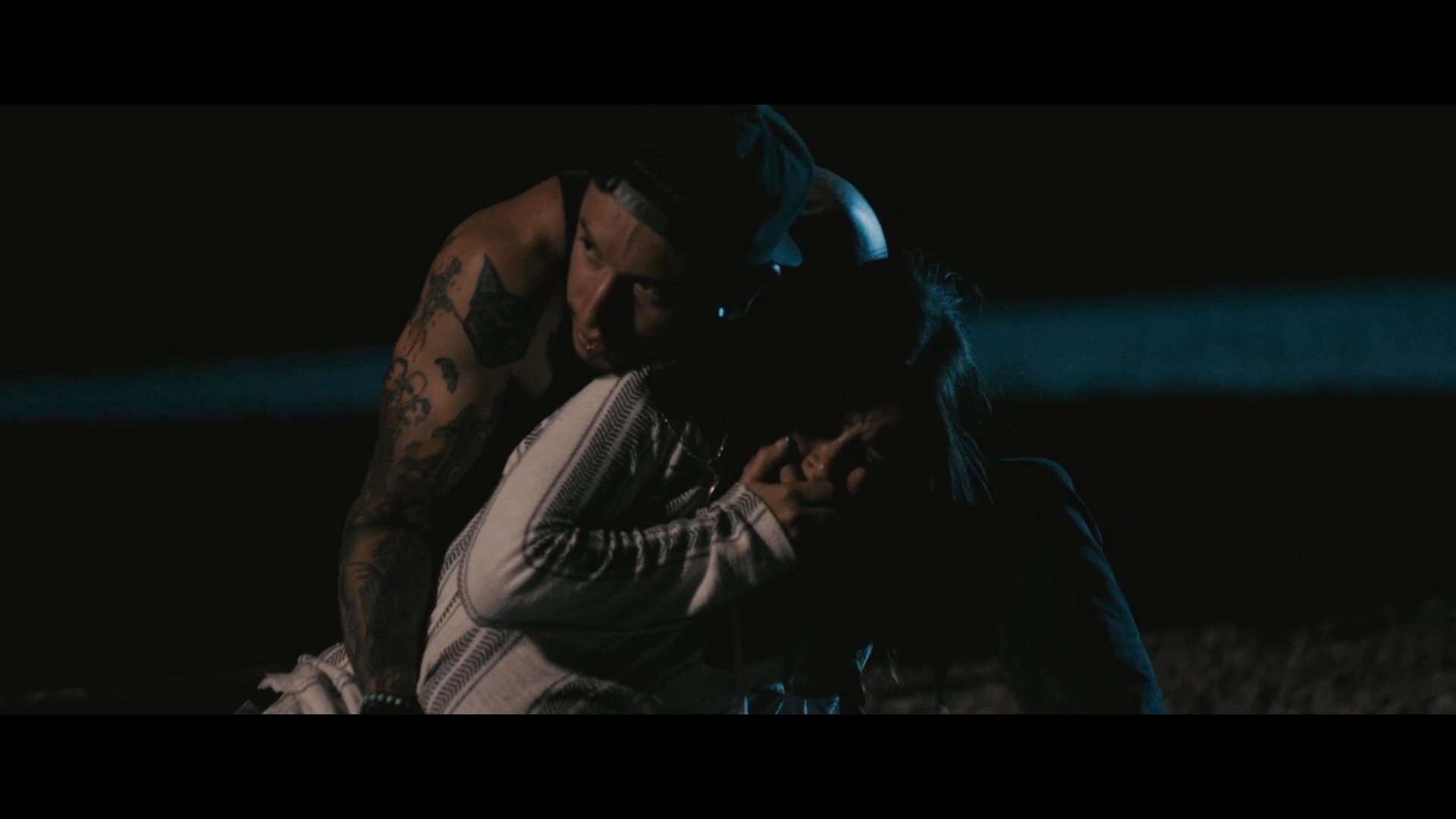 Two individuals engage in a tense, physical struggle outdoors under low-light conditions. The interaction escalates as one person forcibly restrains the other on the ground.