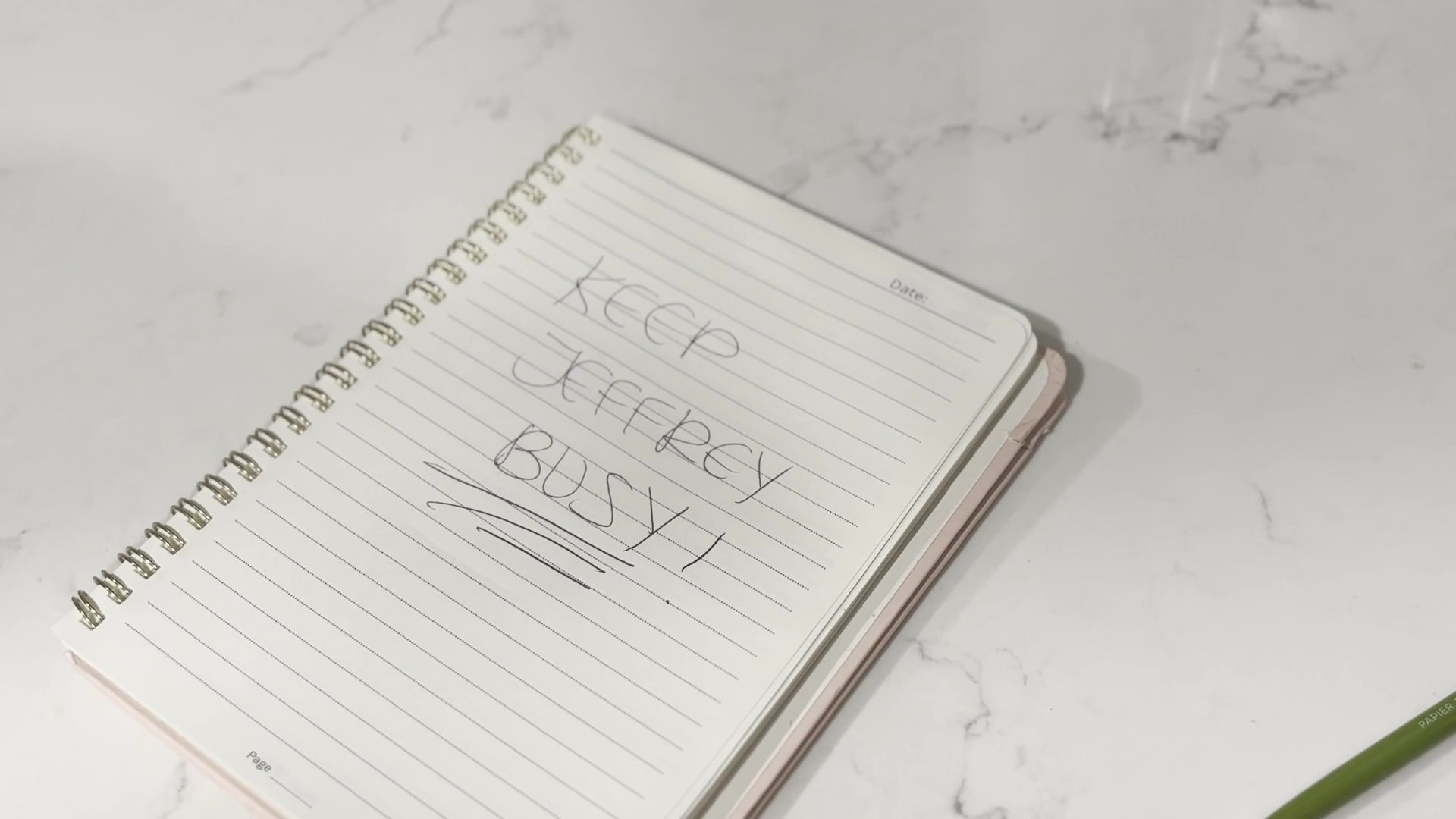 An individual handwrites a list of tasks in a notebook with the heading 'Keep Jeffrey Busy' before turning to address a group of people gathered in a kitchen.