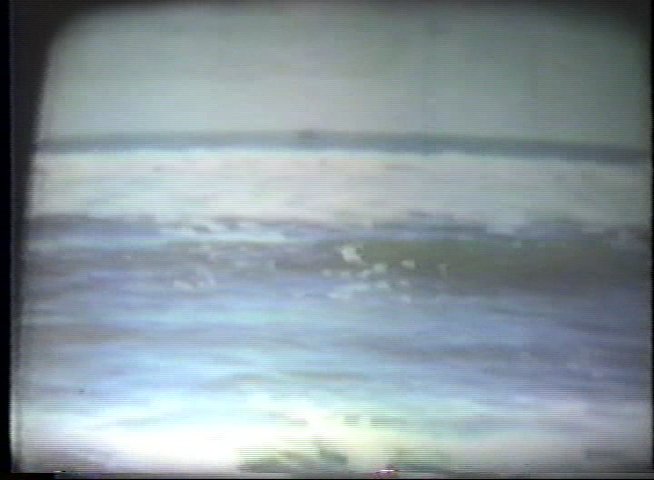 Two individuals provide commentary on archival, black-and-white film sequences of surfers navigating waves near a coastal pier. The footage highlights early twentieth-century surfing techniques in an ocean environment.