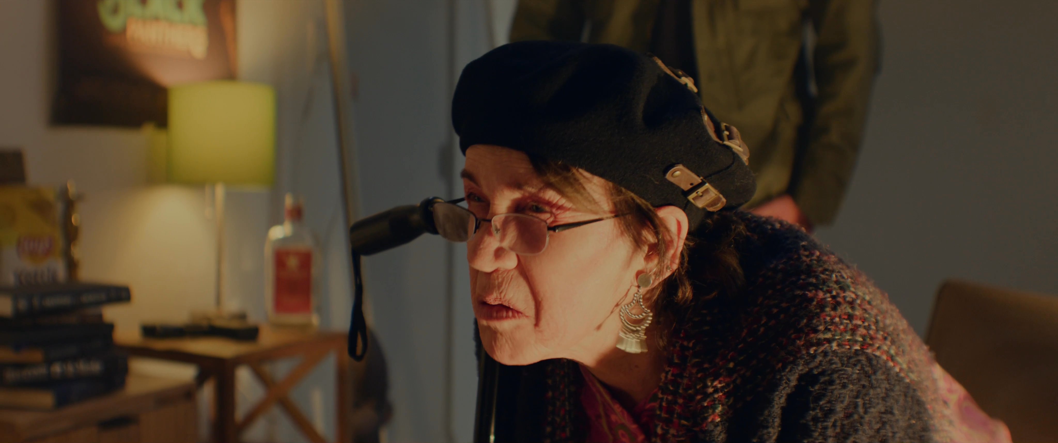 An older woman in a beret engages in a spirited discussion with a younger man within a room decorated with political and revolutionary posters. Their interaction focuses on a direct and intense exchange of viewpoints.