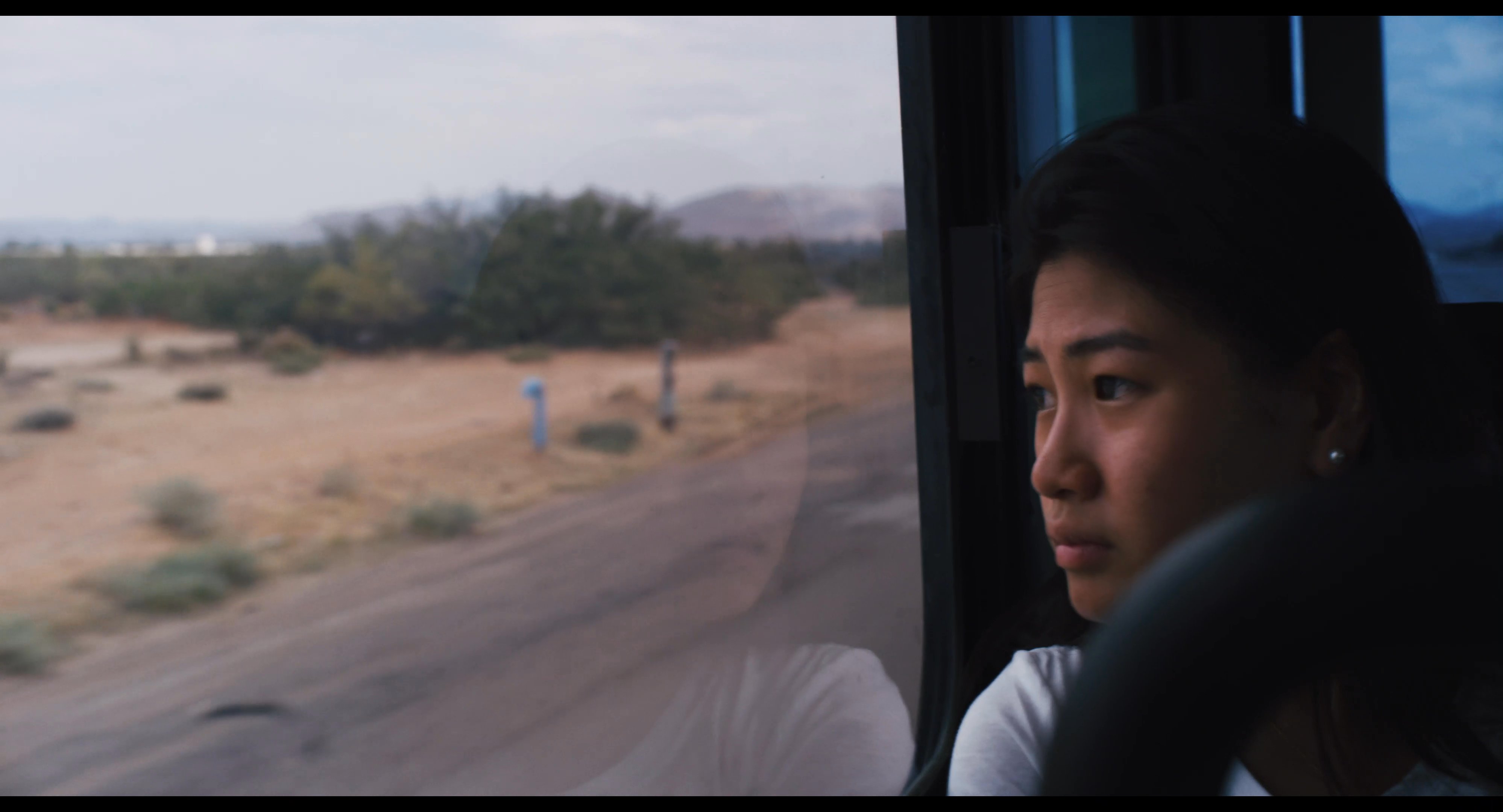 A passenger gazes out the window of a moving bus as it travels along a rural highway past a desert landscape with a prominent water tower.