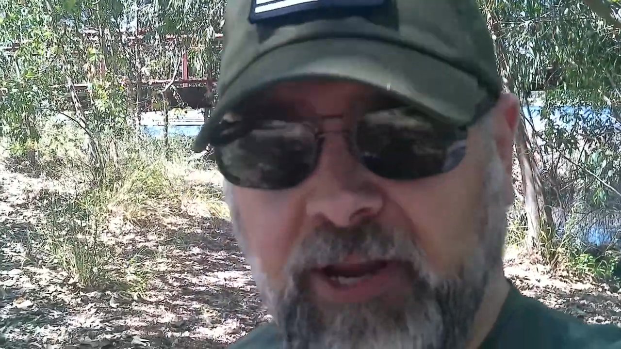 A man wearing sunglasses and a cap records a direct-to-camera commentary while standing in a wooded, sunlit outdoor environment.