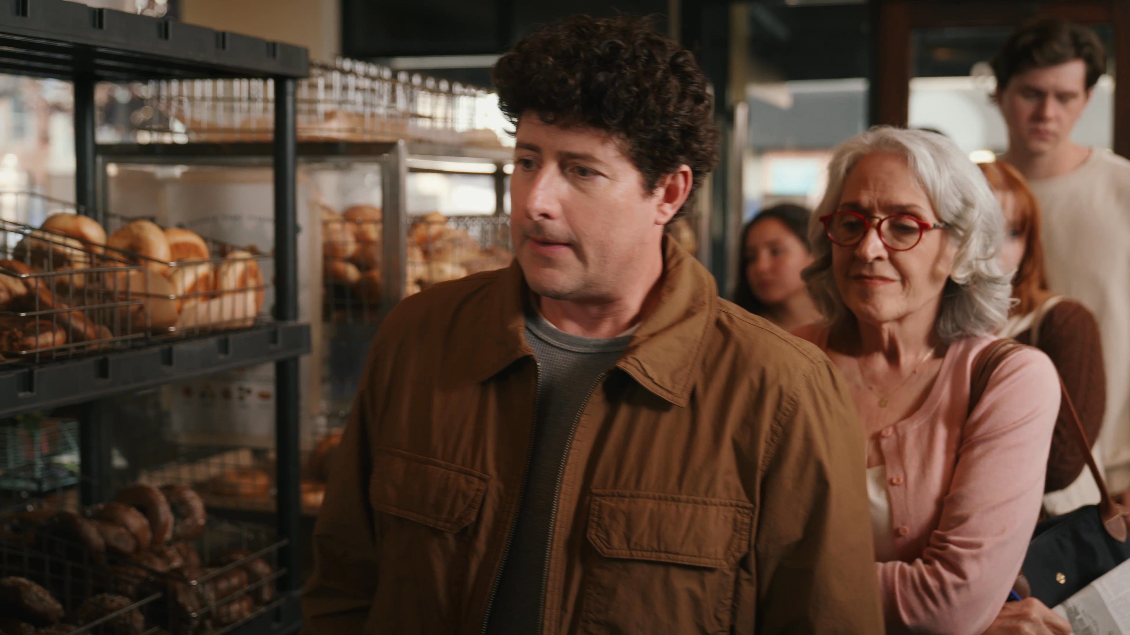 A customer speaks with staff members in a bakery setting amidst racks of bagels. The individuals engage in a brief, professional exchange while waiting in line.