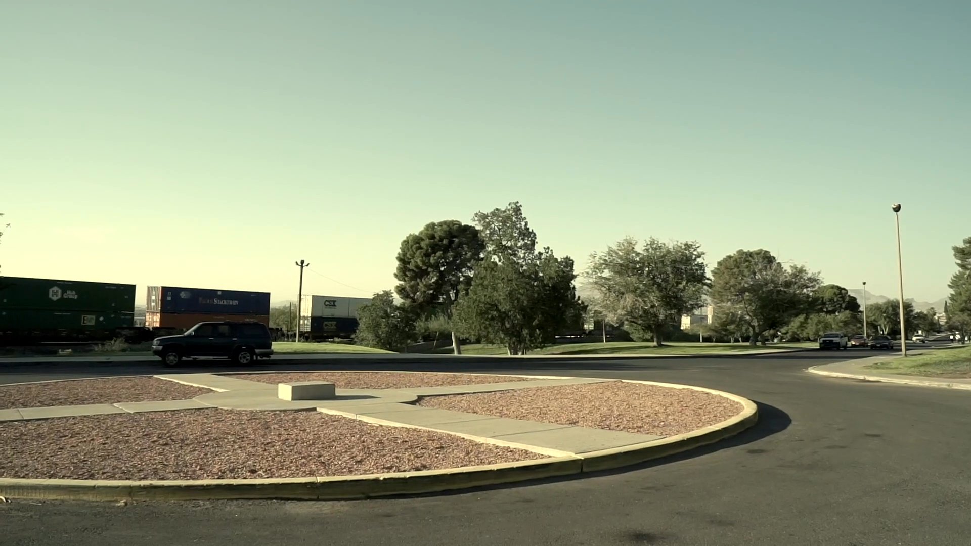 A woman walks across a park at sunset while a man prepares to depart in his vehicle from a nearby parking lot.