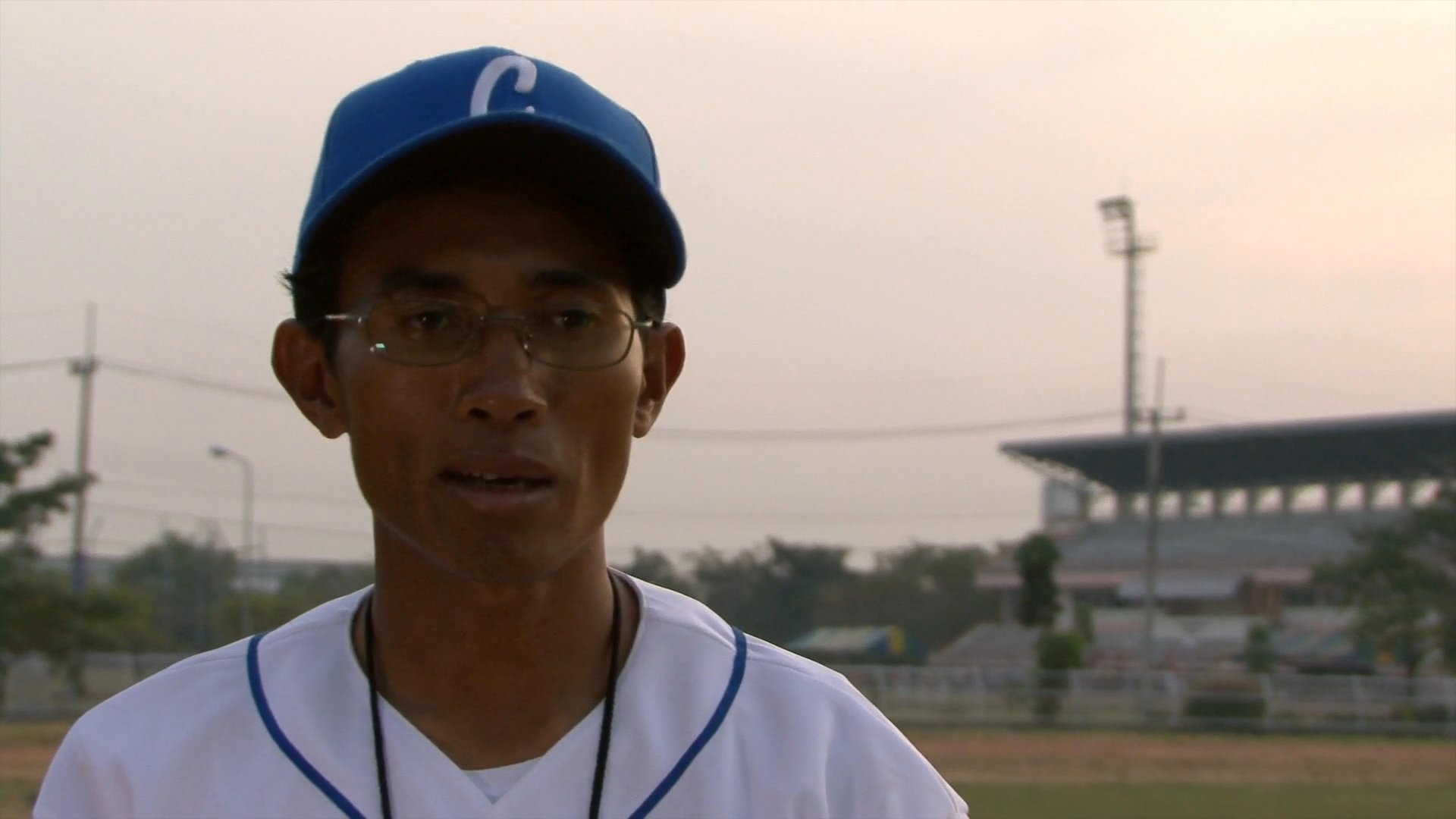 A baseball coach delivers a professional interview while standing on an outdoor field. He speaks directly to the camera, sharing insights regarding his coaching experience and team training.