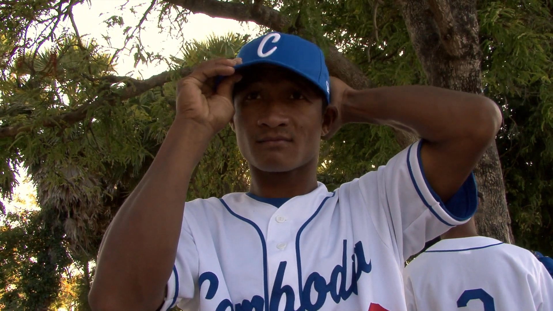 Athletes receive haircuts and prepare in their team uniforms outdoors before a baseball event. The group gathers in a rural setting, finalizing their appearance and equipment for the game.