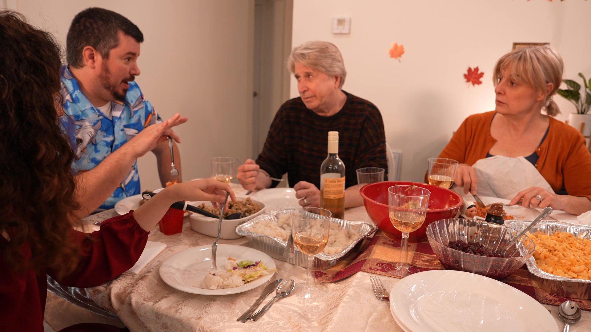 A group of adults sits together at a dining table, engaging in conversation while enjoying a shared meal. The scene captures a casual, intimate social atmosphere within a home environment.