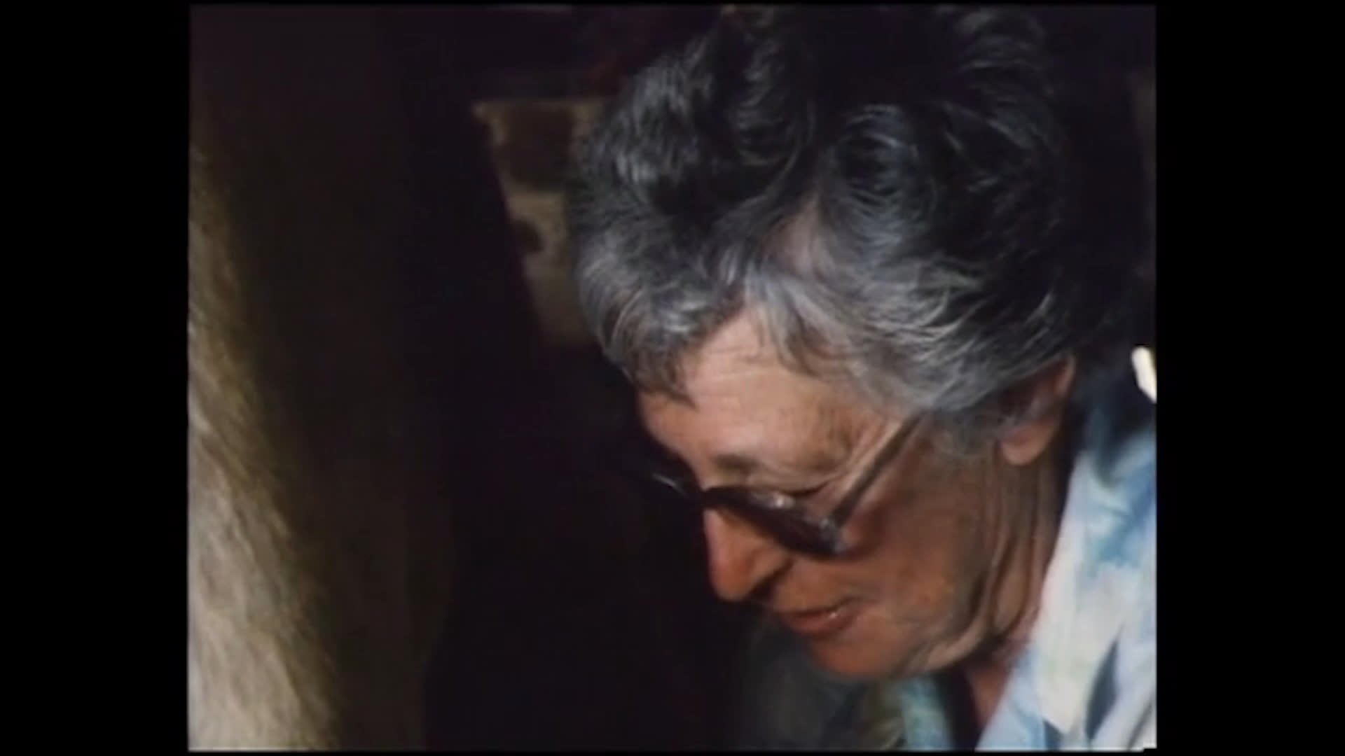 An older woman sits beside a cow in a dim barn, milking as she recalls rising early for farm chores and avoiding cooking and dishes. A pig feeds from a shallow trough nearby.