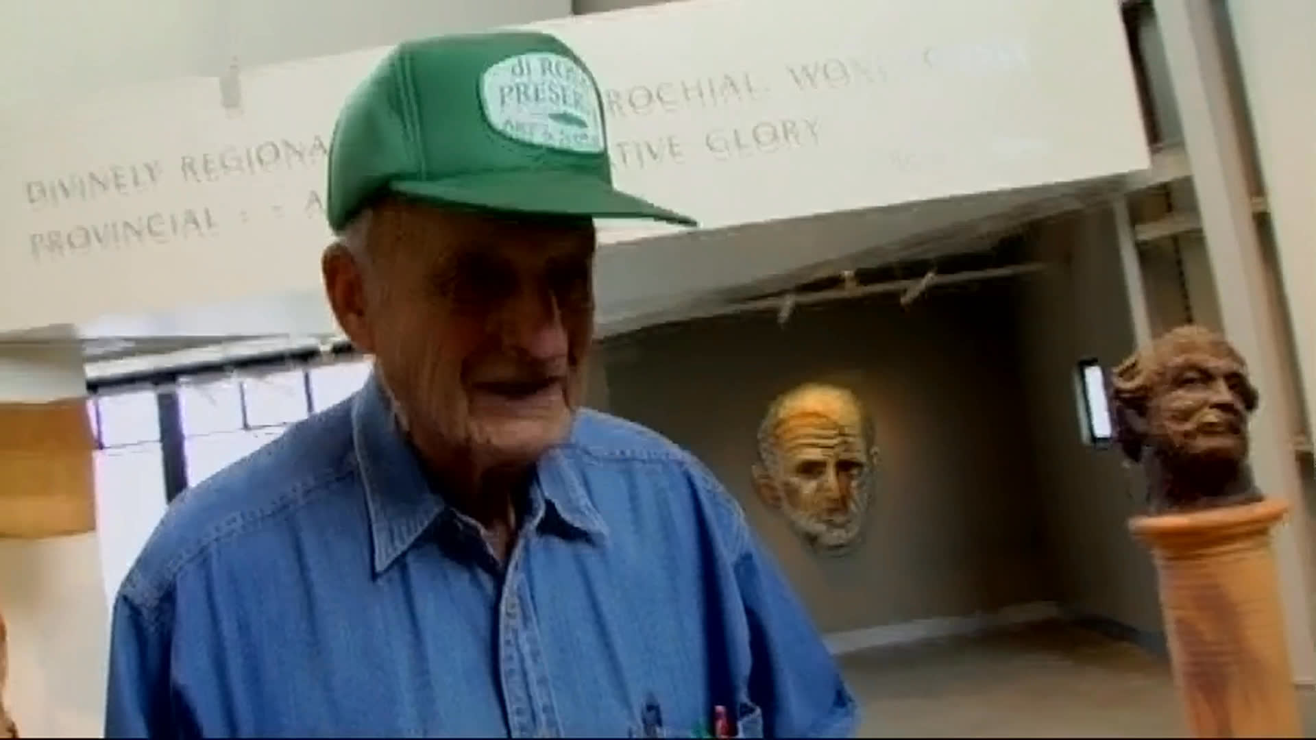 An older man in a green cap discusses an exhibition among portrait busts and other sculptural works, gesturing as he speaks. He explains that a past stroke affects his memory, including his ability to recall artists’ names; brief outdoor views feature a large metal sculpture.