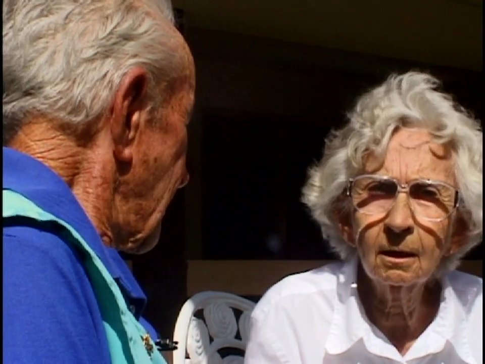 An elderly couple engages in a deliberate, focused conversation while sitting outdoors. The exchange emphasizes personal connection and verbal communication between the two individuals.
