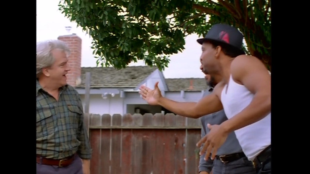 A group of men share a meal at an outdoor table in a residential backyard. They greet arriving acquaintances with friendly handshakes and casual conversation in a relaxed neighborhood setting.