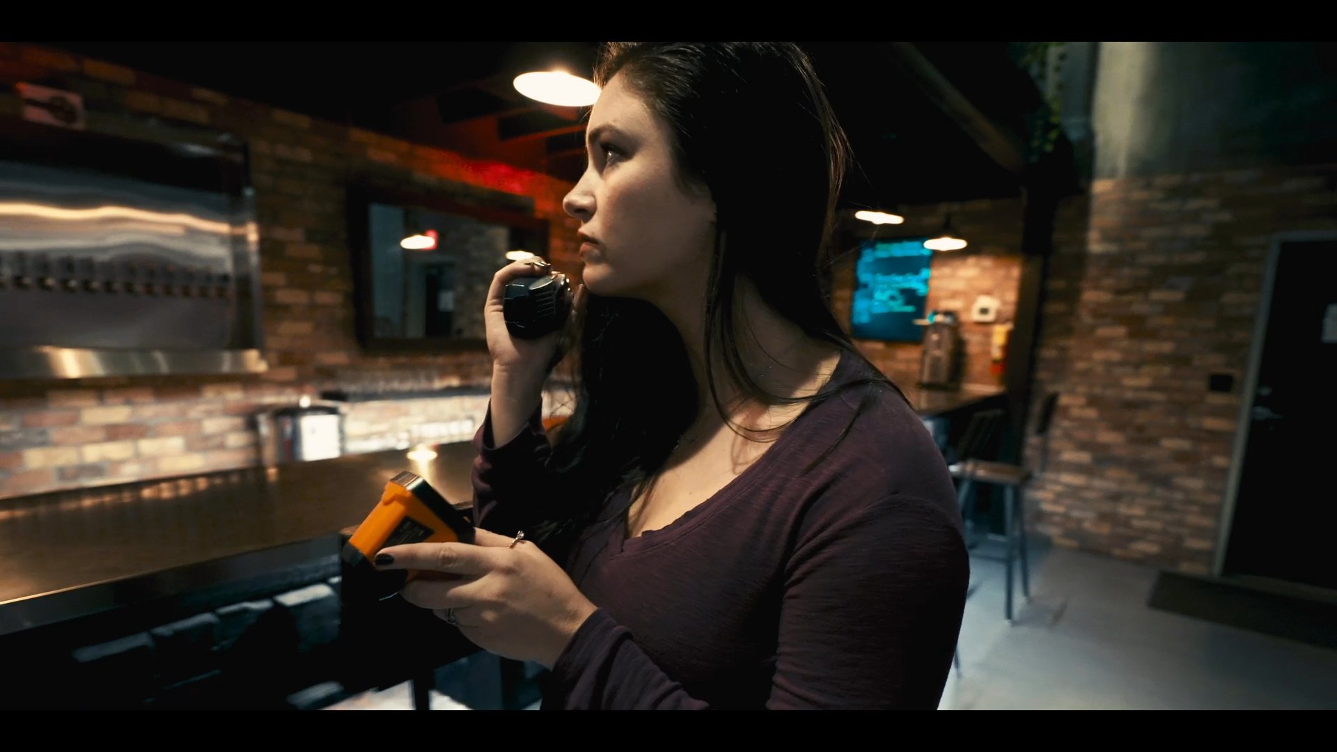 A woman stands in a dimly lit bar, appearing visibly distressed while engaging in a serious phone conversation. She eventually leaves the bar area, moving toward a darker, secluded section of the establishment.