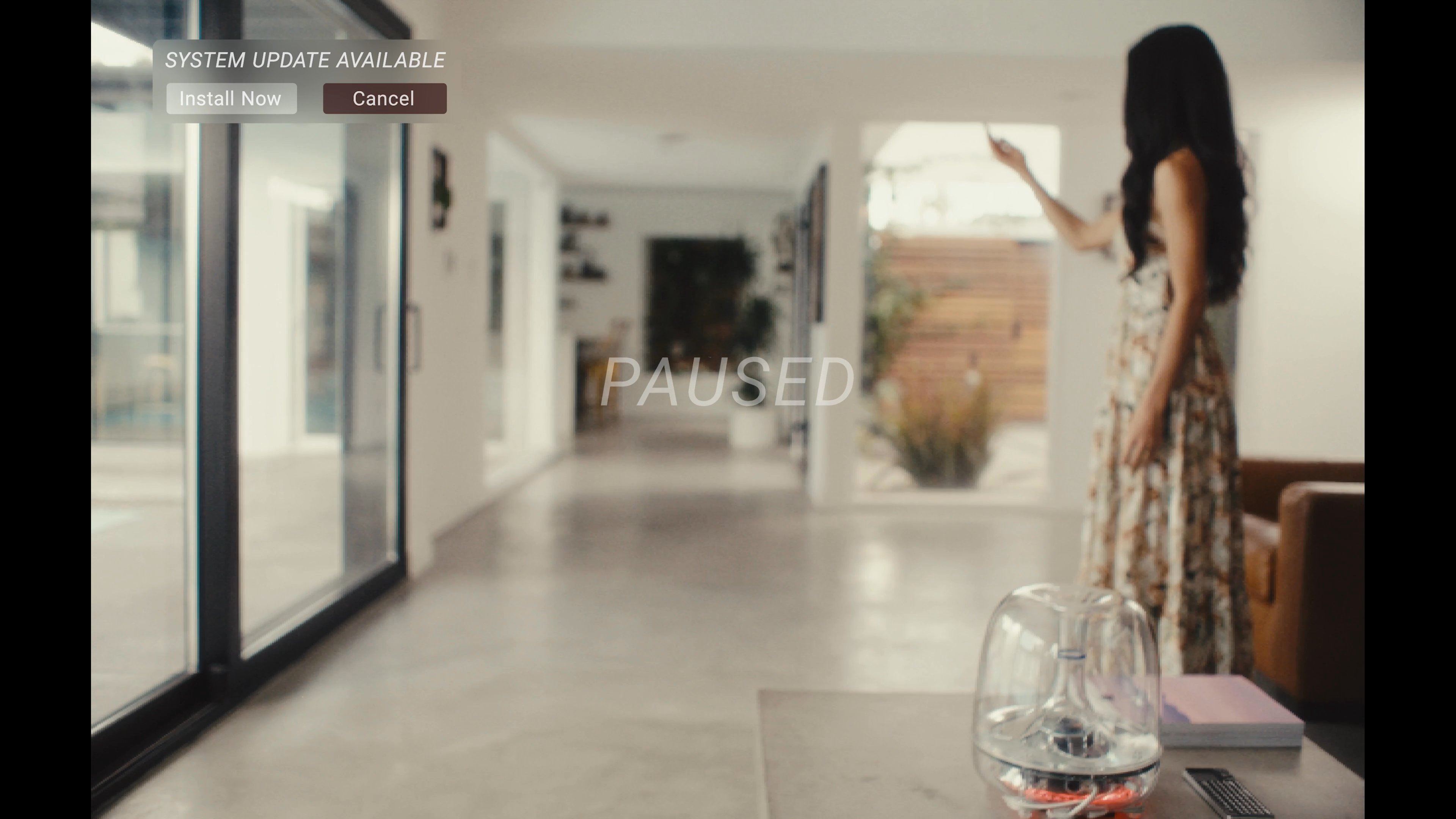 A woman interacts with a digital user interface in a modern living room while a man sits nearby wearing a virtual reality headset. The scene transitions between their shared environment and the digital overlay experienced by the participants.
