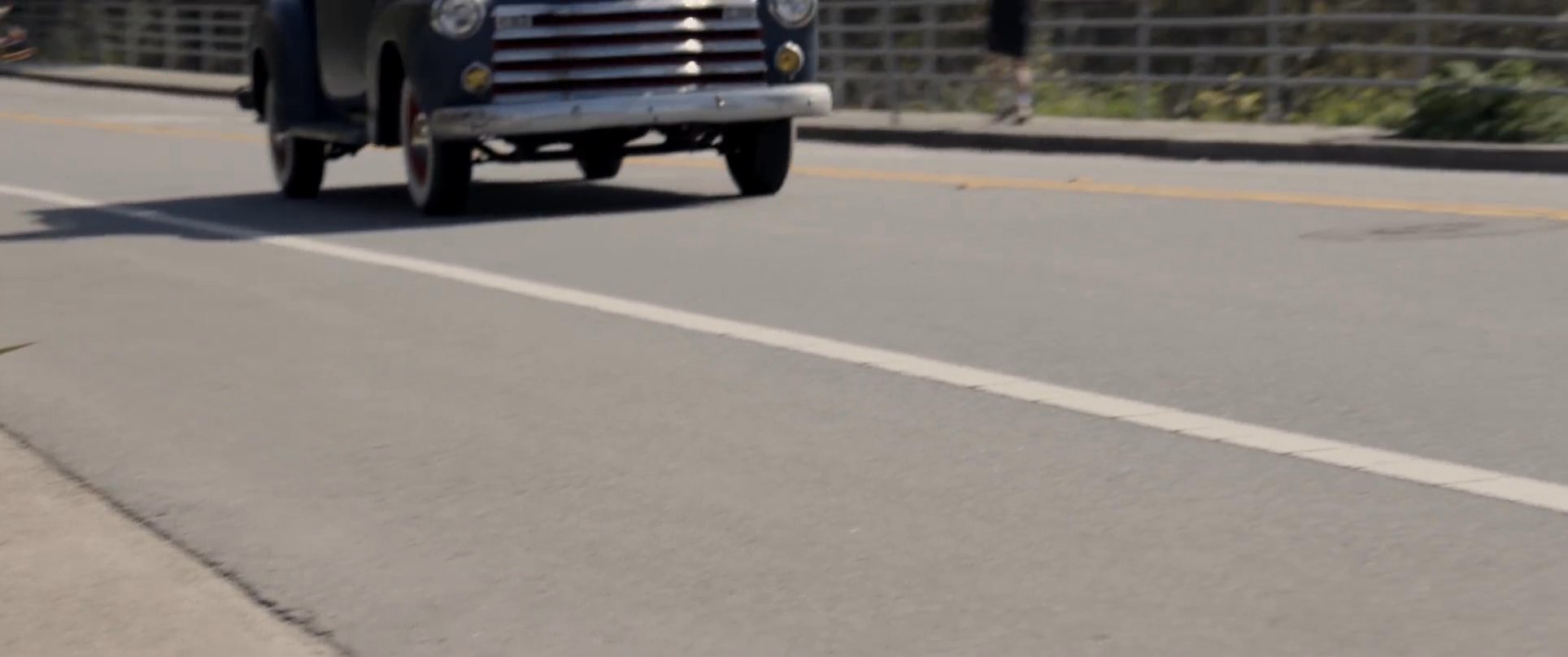 A man drives a classic vintage pickup truck along a scenic coastal highway. The camera captures close-up shots of the driver before transitioning to an aerial view of the vehicle traversing the open road.